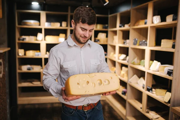Hansome man hold big slice of cheese maasdam in hand. Cheese with big holes. Background of shelves with cheese