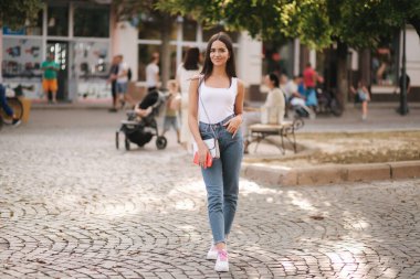 Yaz mevsiminde şehrin merkezinde yürüyen çekici genç bir kadın. Beyaz gömlekli güzel esmer. Gülümse.