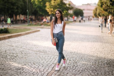 Yaz mevsiminde şehrin merkezinde yürüyen çekici genç bir kadın. Beyaz gömlekli güzel esmer. Gülümse.