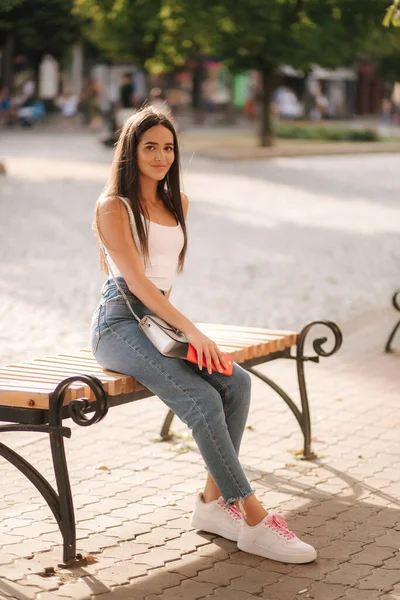 Aantrekkelijke brunette zittend op de bank in het centrum van de stad. mooie jonge vrouw buiten gebruik telefoon — Stockfoto