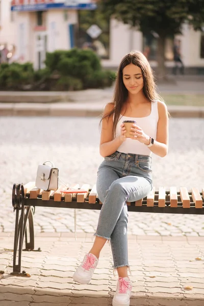 Aantrekkelijke brunette zittend op de bank in het centrum van de stad. Mooie jonge vrouw buiten drinken koffie — Stockfoto