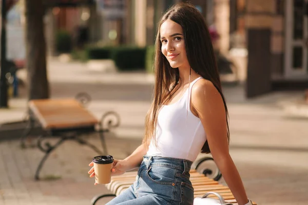 Aantrekkelijke brunette zittend op de bank in het centrum van de stad. Mooie jonge vrouw buiten drinken koffie — Stockfoto