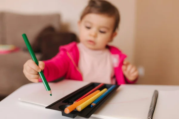 Gadis kecil yang bahagia duduk di meja dan memikirkan sesuatu. Gadis kecil menggunakan pensil untuk menggambar di atas kertas putih di rumah — Stok Foto