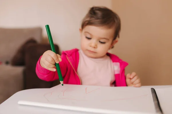 La niña feliz se sienta en la mesa y grita algo. Niña usar lápiz para dibujar en papel blanco en casa —  Fotos de Stock