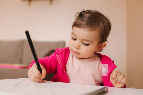 Gelukkige kleine meid zit aan tafel en wthire iets. Klein meisje gebruik potlood voor het tekenen op wit papier thuis — Stockfoto