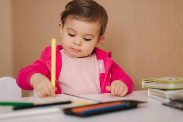 Gelukkige kleine meid zit aan tafel en wthire iets. Klein meisje gebruik potlood voor het tekenen op wit papier thuis — Stockfoto