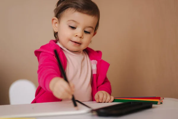 La niña feliz se sienta en la mesa y grita algo. Niña usar lápiz para dibujar en papel blanco en casa —  Fotos de Stock