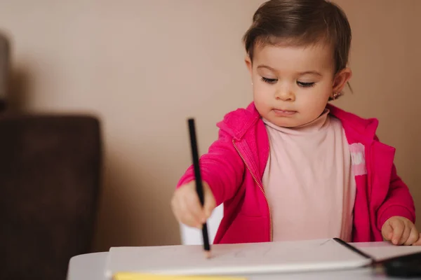 Gelukkige kleine meid zit aan tafel en wthire iets. Klein meisje gebruik potlood voor het tekenen op wit papier thuis — Stockfoto