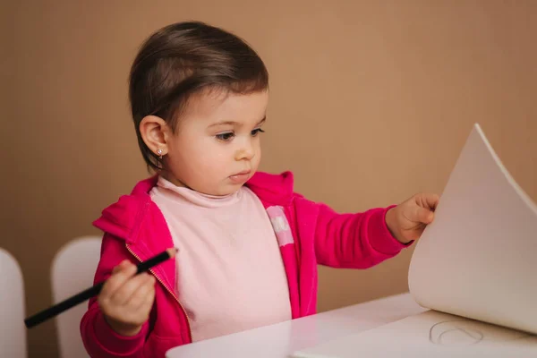 Gelukkige kleine meid zit aan tafel en wthire iets. Klein meisje gebruik potlood voor het tekenen op wit papier thuis — Stockfoto