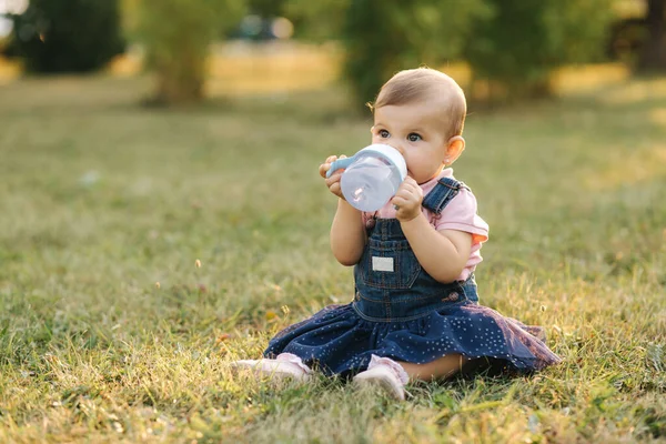Il bambino beve acqua dal biberon fuori nel parco. Bambino gir sedersi sull'erba — Foto Stock