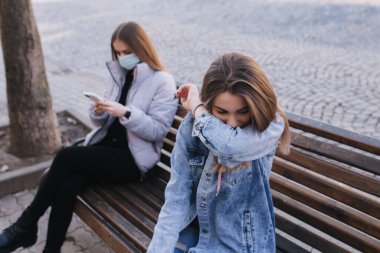 Maskeli kız. Coronavirus teması. Şehirdeki kadınlar karantina sırasında kürsüde koruyucu maskesiz oturuyorlar. Kadın hapşırığı ve öksürüğü