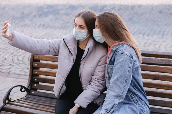 Dziewczyna w maskach. Motyw koronawirusa. Kobiety w mieście siedzą na ławce w masce ochronnej i robią selfie na smartfonie, podczas kwarantanny — Zdjęcie stockowe