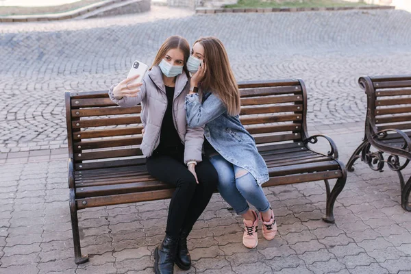 Mädchen in Masken. Coronavirus Thema. Frauen in der Stadt sitzen in Schutzmaske auf der Bank und machen während der Quarantäne ein Selfie mit dem Smartphone — Stockfoto