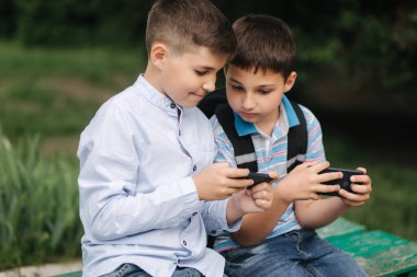 İki çocuk bankta oturuyor ve online oyunlar oynuyorlar. Sırt çantalı bir çocuk. Genç çocuklar telefonlarını kullanır. Karantina