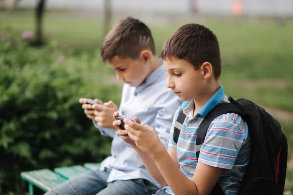 Två pojkar sitter på bänken och spelar onlinespel. En pojke med ryggsäck. Unga pojkar använder sina telefoner. Karantän — Stockfoto
