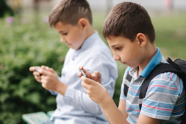 两个男孩坐在长椅上玩网络游戏。一个背着背包的男孩男孩子们用他们的电话。检疫 — 图库照片