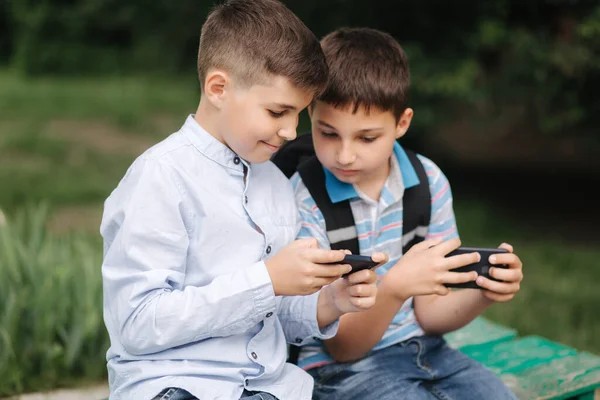 Dva chlapci sedí na lavičce a hrát online hry. Jeden kluk s batohem. Mladí kluci používají telefony. Karanténa — Stock fotografie