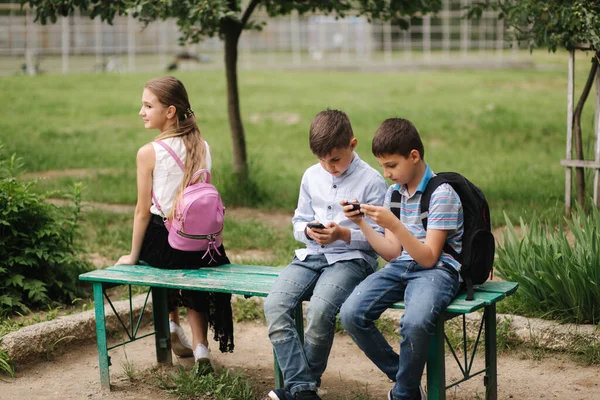 2人の男の子と女の子は学校の休み中に携帯電話を使う。かわいい男の子がベンチに座って、オンラインゲーム — ストック写真