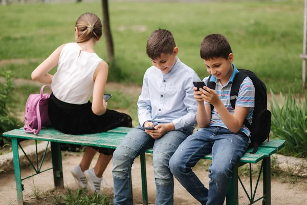 2人の男の子と女の子は学校の休み中に携帯電話を使う。かわいい男の子がベンチに座って、オンラインゲーム — ストック写真