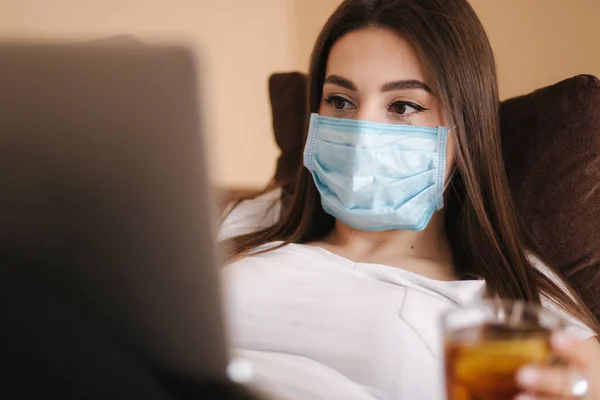 Close up of woman in protective mask have a rest at home during Coronavirus. Woman drinking tea. Female self isolated at home in room. Stay Home