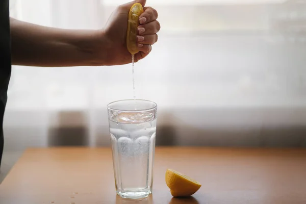 A mão feminina aperta o suco de limão no vidro com a água. Água com limão. Bebida fresca em quarentena — Fotografia de Stock