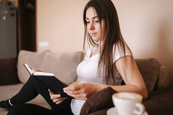 Young attractive woman read the book at home. Quarantine time. Woman drink cappuchino and fill the notebook