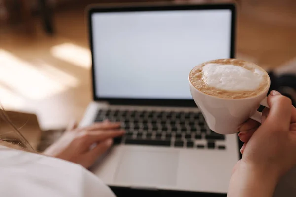 First person view of female working at home. Woman work on laptop and drink cappuccino. Mock up, white screen. Quarantine time