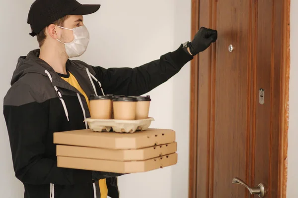 Muž doručuje pizzu a kávu zákazníkovi v masce a rukavicích. Koronavirové téma. Muž klepe na dveře — Stock fotografie