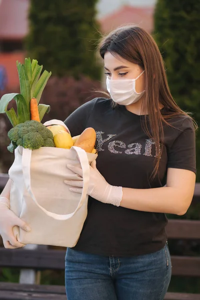 Mladá žena v masce a rukavicích drží tašku s čerstvým jídlem, zeleninovým ovocem a chlebem. Žena po jídle. Chraň se. Motiv koronaviru — Stock fotografie