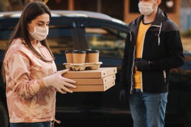 Koruyucu maskeli ve tıbbi maskeli genç kadın dışarıdaki teslimatçıdan emir alıyor. Yemek karantinaya alındı. Kurye müşteri için pizza ve kahve getiriyor.