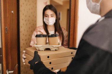 Maskeli ve eldivenli müşterilere pizza ve kahve getiren adam. Kadın kapıyı aç ve siparişi al. Coronavirus teması