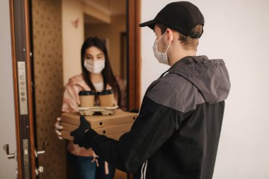 Maskeli ve eldivenli müşterilere pizza ve kahve getiren adam. Kadın kapıyı aç ve siparişi al. Coronavirus teması