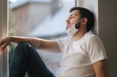 Genç adam pencere pervazına oturur ve koruyucu maskeyi çenesinden aşağı çeker ve pencereden dışarı bakar. Karantinada evde tek başına. Coronavirus teması. Evde kal.
