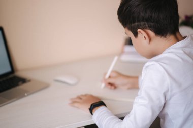 Öğrenci çocuk odasında, masasında ders çalışıyor. Çocuk dizüstü bilgisayar kullanıyor ve deftere yazı yazıyor. Kitaplar ve tablet masanın üstünde. Qurantine sırasında evde çalış
