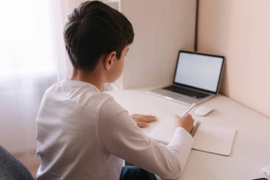 Öğrenci çocuk odasında, masasında ders çalışıyor. Çocuk dizüstü bilgisayar kullanıyor ve deftere yazı yazıyor. Kitaplar ve tablet masanın üstünde. Qurantine sırasında evde çalış