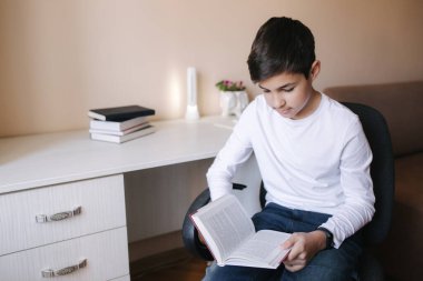 Genç çocuk masanın yanında oturuyor ve kitabı okuyor. Karantina sırasında evde çalış