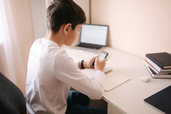 Школьник учится за столом в своей комнате. Мальчик использует ноутбук и смартфон для записи в блокнот. Книги и планшеты на столе. Обучение дома во время qurantine — стоковое фото