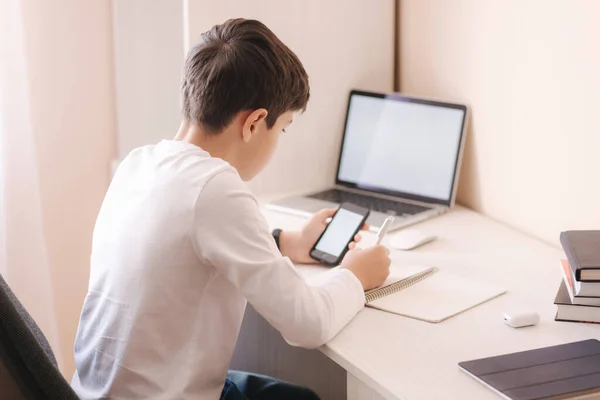 Estudiante de escuela en el escritorio de su habitación. El muchacho usa el portátil y el smartphone para escribir en el portátil. Libros y tabletas sobre la mesa. Estudiar en casa durante la cuarentena — Foto de Stock