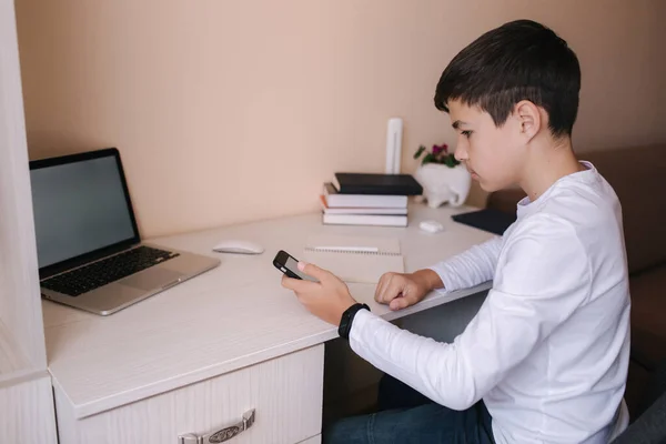 Estudiante de escuela en el escritorio de su habitación. El muchacho usa el portátil y el smartphone para escribir en el portátil. Libros y tabletas sobre la mesa. Estudia en casa durante la cuarentena. Prepárate. — Foto de Stock