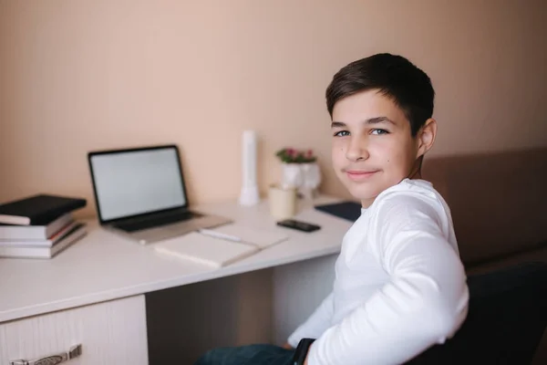 Щасливий хлопчик сидить за столом і вчиться вдома. Хлопчик навчається онлайн під час карантину. Фон ноутбука та книг — стокове фото