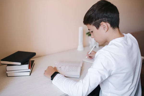Ο νεαρός που κάθεται στο γραφείο διαβάζει το βιβλίο και γράφει στο σημειωματάριο. Μελέτη στο σπίτι κατά τη διάρκεια καραντίνας — Φωτογραφία Αρχείου