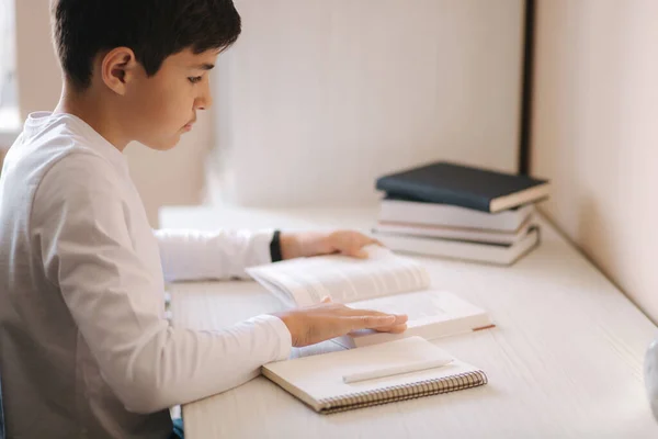 Jonge jongen zit aan het bureau lees het boek en whright neer in notebook. Studie thuis tijdens quarantaine — Stockfoto