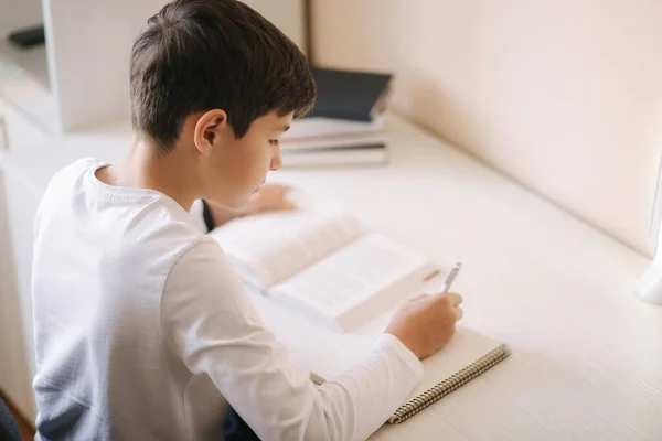 Jonge jongen aan het bureau las het boek en schreef het op in het notitieboekje. Studie thuis tijdens quarantaine — Stockfoto