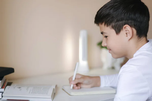 Ein kleiner Junge, der am Schreibtisch sitzt, liest das Buch und schreibt es in ein Notizbuch. Während der Quarantäne zu Hause studieren — Stockfoto