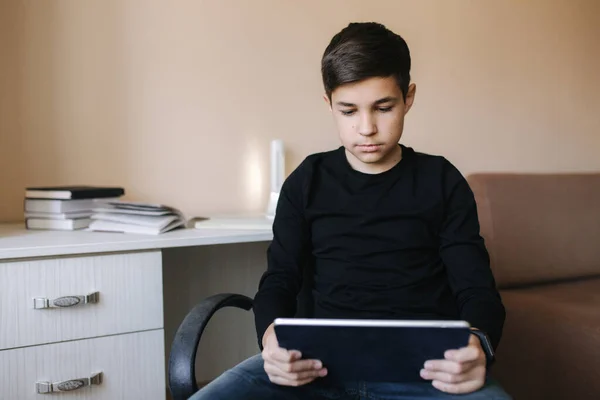 O adolescente em casa senta-se à mesa e usa o comprimido durante o intervalo. Jovem menino jogar jogos online no tablet. Backgroung de mesa com livros — Fotografia de Stock