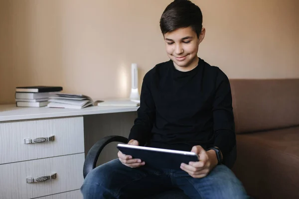 O adolescente em casa senta-se à mesa e usa o comprimido durante o intervalo. Jovem menino jogar jogos online no tablet. Backgroung de mesa com livros — Fotografia de Stock