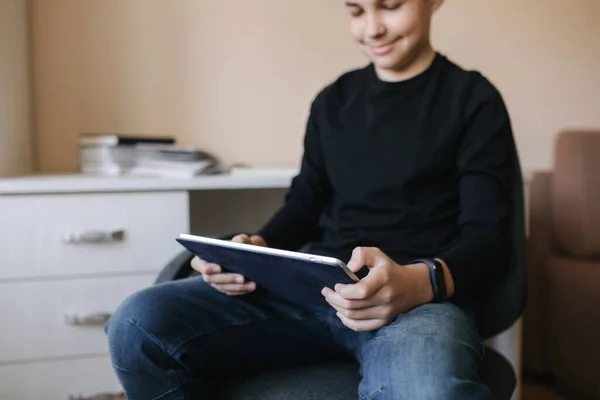 家里的少年坐在桌旁，休息时用平板电脑。小男孩在平板电脑上玩网络游戏。书桌的背景图 — 图库照片