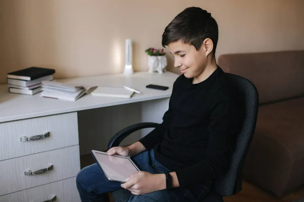 O adolescente em casa senta-se à mesa e usa o comprimido durante o intervalo. Jovem menino jogar jogos online no tablet. Backgroung de mesa com livros — Fotografia de Stock