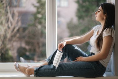 Genç bir kadın pencere kenarında oturuyor ve koruyucu maskeyi elinde tutuyor. Kadın ve pencereden dışarı bak. Karantinada evde tek başına. Coronavirus teması. Evde kal.