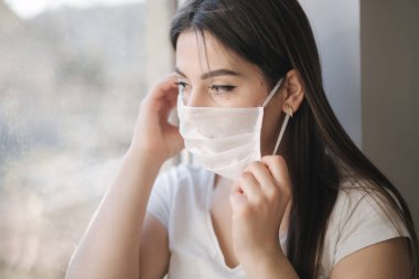 Genç bir kadın pencere pervazında ve koruyucu maskeyle oturur ve pencereden dışarı bakar. Karantinada evde tek başına. Coronavirus teması. Evde kal.